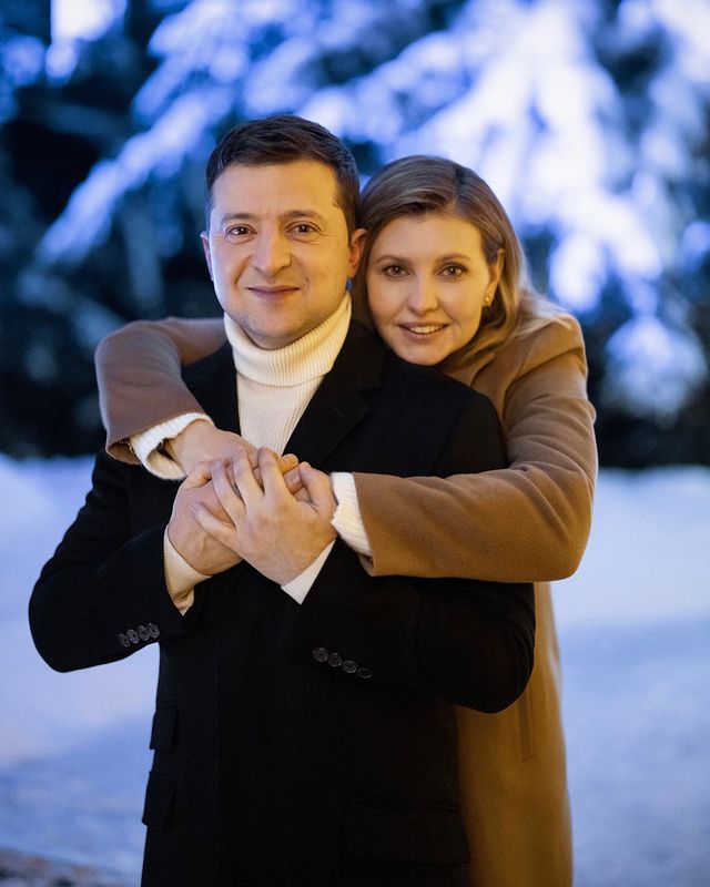 Olena Zelenska in a brown coat hugging her husband, Volodymyr Zelenskyy, in a white high neck and black winter coat.